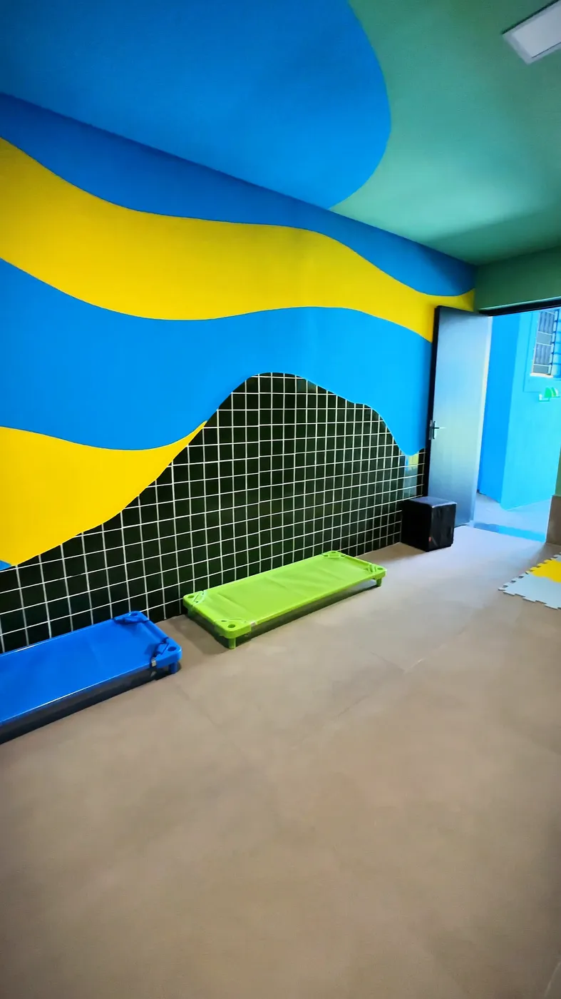 Ambiente de descanso com azulejo verde, mural em onda azul e amarelo e caminhas elevadas